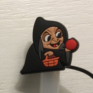 May include: A black cartoon witch figure with a red apple and orange basket. The witch has a tan face, a black hood, and is holding a red apple. The figure is attached to a white charging cable.