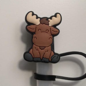 May include: A brown moose with large antlers sits on a black silicone straw holder. The moose has a cute expression and is wearing a black and gray hat.