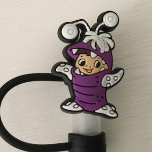May include: Purple and white character charm with a cartoon monster design. The character is wearing a purple costume with white eyes and a white smile. The charm is attached to a black elastic band.