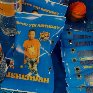 May include: Blue party favor bags with a photo of a boy in an orange shirt, the text "HAPPY 7TH BIRTHDAY", and "NERF" logo. Hershey's chocolate bars and a cupcake with orange frosting are also visible.
