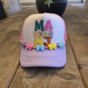 May include: A light purple trucker hat with the word "MA HAT" in colorful, sequined letters. The hat has a white front panel, pink mesh sides, and is decorated with small bows in various colors.