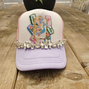 May include: A trucker hat with a lavender bill, white crown, and pink mesh back. The crown features an embroidered design with a peacock, flowers, and decorative elements. The bill is adorned with a row of clear, rectangular rhinestones.