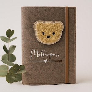 Könnte beinhalten: Eine braune Filzhülle mit einem Teddybär-Applikation und dem Wort "Mutterpass" in Schrift. Die Hülle hat ein braunes Gummiband und ein kleines Herzsymbol. Der Teddybär ist beige mit schwarzen Details.