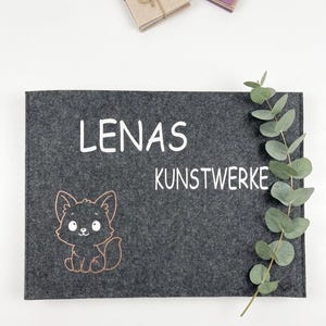 Könnte beinhalten: Eine dunkelgraue Filzmatte mit dem Text "LENAS KUNSTWERKE" in Weiß. Eine Cartoon-Fuchs-Illustration befindet sich unten links. Eukalyptusblätter befinden sich auf der rechten Seite. Zwei kleine Bündel sind im Hintergrund.