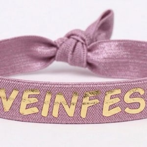 Könnte beinhalten: Ein mauvefarbenes Haargummi mit dem goldfarbenen Schriftzug "WEINFEST". Das Band besteht aus einem strukturierten Stoff und ist oben verknotet. Dieses Haaraccessoire dient dazu, das Haar zu fixieren.