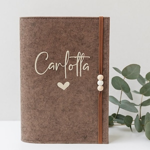 Könnte beinhalten: Ein braunes Filzjournal mit dem Namen "Carlotta" in Schreibschrift und einem kleinen Herz. Ein braunes Gummiband mit Holzperlen sichert das Journal. Eukalyptusblätter sind im Hintergrund.