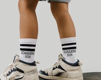 Puede incluir: Una persona con calcetines blancos con rayas negras y el texto "SCHULKIND 2026", combinados con zapatillas altas color crema con detalles azul marino. La persona está de pie sobre una patineta, usando pantalones cortos de mezclilla.