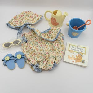 May include: A doll-sized summer set including a cream-coloured romper and sun hat with a multi-coloured beach print, blue sandals with sun charms, yellow watering can, blue bucket with a shovel, white sunglasses, and a Bitty Bear's Summer Fun book.