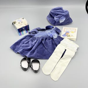 May include: A doll outfit set including a purple velvet dress with a matching hat, both featuring blue bows. The set includes white tights with lace trim, black Mary Jane shoes, a small white plush cat in a striped box, and a book titled Billy Bear's Winter Fun.