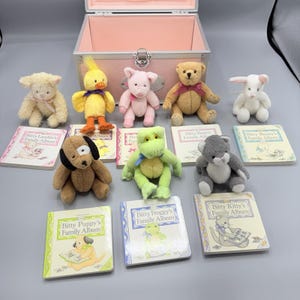 May include: A collection of plush animal toys including a lamb, duck, pig, bear, rabbit, dog, frog, and cat, each paired with a corresponding Bitty Family Album book. A light pink metal storage case with a silver latch is positioned behind the toys.
