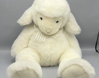 May include: A plush stuffed animal in the shape of a lamb, covered in soft, off-white faux fur. The lamb features long, floppy ears, dark eyes, and a cream-colored ribbon tied in a bow around its neck with thin, vertical white stripes.