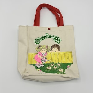 May include: Cream-colored canvas tote bag with red handles. The bag features a colorful graphic of Cabbage Patch Kids watering cabbages. The text "Cabbage Patch Kids" is printed above the graphic. The bag has a button closure.