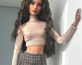 Puede incluir: Una muñeca con el pelo largo y ondulado marrón que lleva un top corto de cuello alto beige y una falda de cuadros marrones.