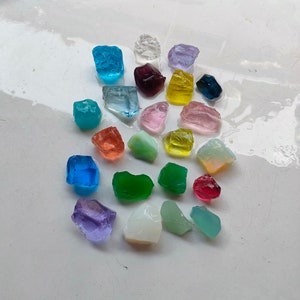 May include: Assortment of colorful glass stones in various shapes and sizes. The stones are arranged on a white surface.