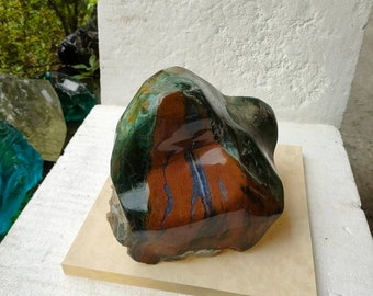 May include: A large, irregularly shaped, polished stone with green, brown, and purple hues. The stone is sitting on a light beige square base.
