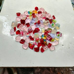 Puede incluir: Una colección de pequeñas piezas de vidrio de colores en tonos rojos, rosas y transparentes. Las piezas están dispuestas en círculo sobre una superficie blanca.