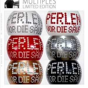 May include: Five round ornaments with the text "PERLEN VOR DIE SAU" spelled out in white pearls on a variety of colored backgrounds. The backgrounds are silver, red, gold, black, and white.