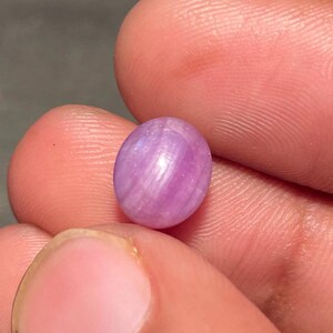 May include: A small, oval-shaped, pink gemstone with a subtle, swirling pattern. The gem is held between two fingers, showcasing its polished surface and natural color variations. The gemstone appears to be a cabochon, suitable for jewelry making.
