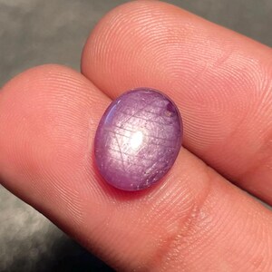 May include: A polished, oval-shaped, purplish-pink gemstone. The gem has a smooth surface with a subtle, shimmering effect. The stone is held between fingers, showcasing its colour and shape.