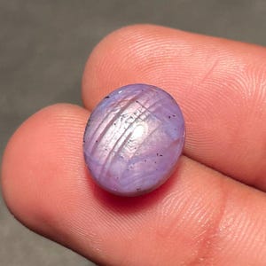 May include: Oval-shaped, lavender gemstone with a star effect. The polished surface displays a cross-hatched pattern of light and dark lines. The gemstone is held between fingers, showcasing its colour and texture.