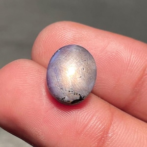 May include: An oval gemstone with a mix of purple, silver, and gold hues. The polished surface reflects light, creating a shimmering effect. The gemstone is held between two fingers, showcasing its size and shape.
