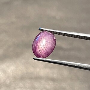 May include: A polished, oval-shaped, purplish-red gemstone is held by tweezers. The gem has a star-like effect, with lines radiating from the centre. The background is a dark, neutral colour.