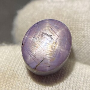 May include: A polished, oval-shaped lavender star sapphire gemstone featuring a distinct six-rayed star pattern visible on its smooth, cabochon-cut surface. The stone displays subtle internal inclusions and a soft, light purple hue throughout its body.