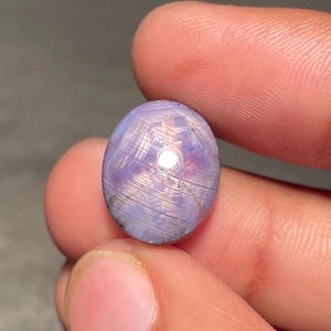 Puede incluir: Gema ovalada de color lavanda con efecto de estrella. La superficie pulida refleja la luz, creando una apariencia brillante. La gema se sostiene entre los dedos, mostrando su tamaño y color.