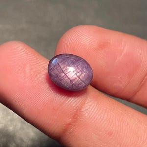 May include: A small, oval, purplish-pink gemstone with a star pattern. The gem is held between two fingers, with tweezers in the background. The stone's surface reflects light, creating a star effect. The gem is likely a star sapphire.