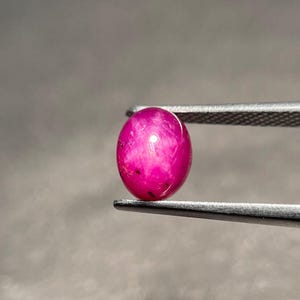 Könnte beinhalten: Ein leuchtender, oval geformter Cabochon-Rubin mit einem tiefen magentaroten Farbton und einer glatten, polierten Oberfläche. Der Edelstein wird sicher zwischen den gezackten Metallspitzen einer silberfarbenen Pinzette gehalten.