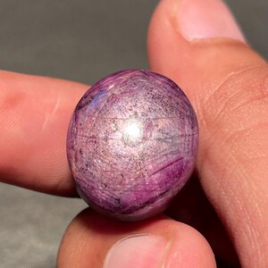 Könnte beinhalten: Ein kugelförmiger, polierter Edelstein in tiefem Magenta, der ein subtiles Lichtspiel auf seiner Oberfläche aufweist. Die Farbe des Steins variiert von einem dunklen, fast schwärzlichen Lila bis zu einem leuchtenden Rosa, mit sichtbaren inneren Mustern. Der Edelstein wird zwischen zwei Fingern gehalten.