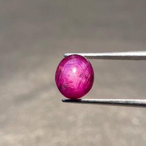 Peut inclure: Une pierre précieuse rubis étoilé de taille cabochon ronde avec une teinte magenta-rouge profonde. La pierre présente un effet d'étoile à six branches distinct centré sur sa surface, maintenue par une pince en métal argenté sur un fond neutre.