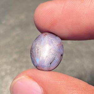 Op de afbeelding: Ovale edelsteen met zilveren, blauwe en zwarte tinten. De gepolijste steen wordt tussen twee vingers gehouden, waardoor het gladde oppervlak en de kleurvariaties worden benadrukt. De edelsteen wordt waarschijnlijk gebruikt voor het maken van sieraden.