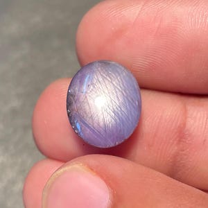 Puede incluir: Gema ovalada pulida, de color lavanda y brillo iridiscente. La piedra muestra un sutil juego de luces, con vetas plateadas y azules en su superficie. La gema se sostiene entre los dedos, mostrando su tamaño y color.