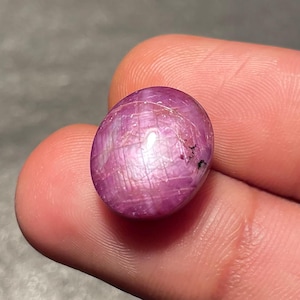 May include: A pinkish-red, oval gemstone with a polished surface. The gem displays internal lines and a subtle sheen. The stone is held between fingers, highlighting its shape and colour. The gem is likely a ruby.