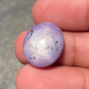 May include: A small, oval gemstone with a star pattern. The stone is a soft lavender colour with white and black accents. The gemstone is held between fingers, showcasing its intricate design and polished surface.