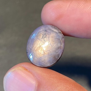May include: An oval, polished gemstone in a light purple shade. The stone displays a delicate star-like design on its surface. The gem is held between two fingers, highlighting its form. The backdrop is a dark, neutral tone.
