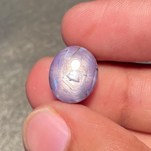 May include: A faceted, oval gemstone in a pale lavender colour. The gem features a star-like pattern on its surface, with blue and white highlights. The stone is held between two fingers, highlighting its size and detail.