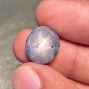 Puede incluir: Una pequeña piedra preciosa redonda y pulida, de color predominantemente gris, con destellos iridiscentes azules y morados. La piedra se sostiene entre los dedos, mostrando su superficie lisa y reflectante. La piedra preciosa parece ser labradorita.