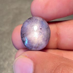 May include: An oval gemstone, polished to a high sheen, displaying lavender and silver tones. The stone is held between two fingers. The gemstone has an iridescent quality and is a natural product.