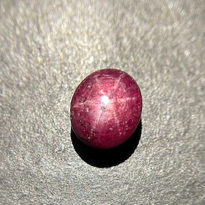 Pode incluir: Uma pedra preciosa de rubi estrela lapidada em cabochão com um tom vermelho magenta profundo. A pedra apresenta um efeito de estrela de seis raios, conhecido como asterismo, centrado na sua superfície lisa, polida e arredondada.