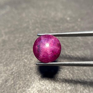Puede incluir: Una piedra preciosa de rubí estrella de corte cabochon redondo con un tono rojo magenta intenso. La piedra muestra un efecto de estrella de seis rayos, conocido como asterismo, centrado en su superficie pulida. Sujeta por unas pinzas metálicas.