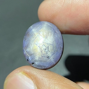 May include: A round, cabochon-cut star sapphire gemstone featuring a light purple and grey hue with a visible six-rayed star effect across its surface. The stone displays a smooth, polished finish and a distinct chatoyancy pattern throughout.