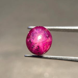 Puede incluir: Una piedra preciosa de rubí estrella de corte cabujón redondo, con un color rojo magenta intenso y un efecto de estrella de seis puntas visible. La gema presenta inclusiones internas en forma de aguja y está sujeta por pinzas de metal plateado.
