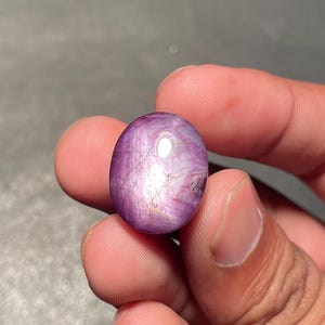 Könnte beinhalten: Oval geschliffener Edelstein mit tiefvioletter Farbe und Sterneneffekt. Der Stein hat eine glatte, reflektierende Oberfläche mit inneren Linien und einem subtilen Lichtspiel. Der Edelstein wird in einer Hand gehalten.