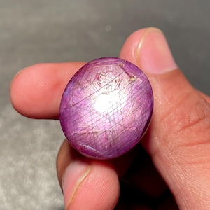 May include: A polished, oval gemstone in a deep magenta colour. The gem exhibits a star-like effect, with lines radiating from a central point. The stone is held between fingers, highlighting its size and colour.