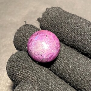 Op de afbeelding: Een ronde, gepolijste edelsteen met een levendige roze tint en een stervormig patroon. De edelsteen wordt vastgehouden door een zwarte gehandschoende hand, die de kleur en het ingewikkelde ontwerp benadrukt. Het oppervlak van de edelsteen reflecteert licht.