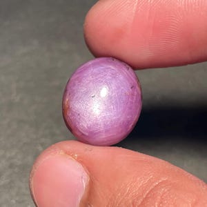 Könnte beinhalten: Ein polierter, oval geformter, rosa Edelstein mit Sterneneffekt. Der Edelstein wird zwischen zwei Fingern gehalten, wodurch seine glatte Oberfläche und Farbvariationen zur Geltung kommen. Der Edelstein scheint ein Rubin zu sein, eine beliebte Wahl für Schmuck.