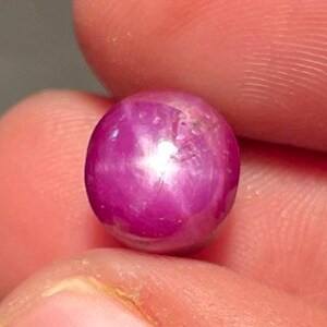 Puede incluir: Una gema de rubí redonda y pulida con efecto estrella. La gema es de color rojo rosado intenso y parece estar sujeta entre los dedos. La superficie del rubí es lisa y reflectante, con un patrón en forma de estrella visible.