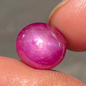 May include: A polished, oval-shaped, ruby gemstone. The gem is a vibrant pinkish-red color with a smooth, reflective surface. The stone is held between two fingers, showcasing its size and shape. The gemstone is a natural, precious stone.